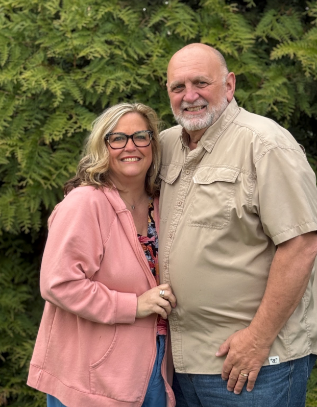 Couple posing outdoors in casual attire against greenery.